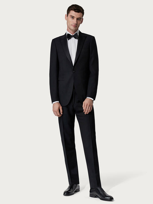 A young man stands against a light gray background wearing a Canali Siena Black Wool Tuxedo With Peak Lapels, a bow tie, and polished black dress shoes.
