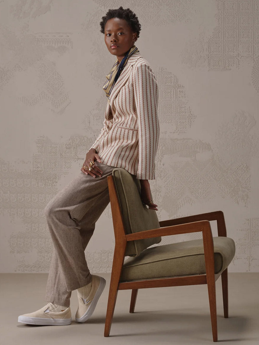 A person wearing the Circolo 1901 Slim Striped Long Jacket in Naturale Knitted, a scarf, beige pants, and sneakers sits sideways on a green cushioned wooden chair near a patterned wall.