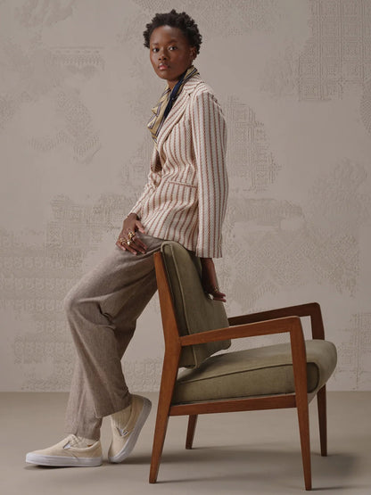 A person wearing the Circolo 1901 Slim Striped Long Jacket in Naturale Knitted, a scarf, beige pants, and sneakers sits sideways on a green cushioned wooden chair near a patterned wall.