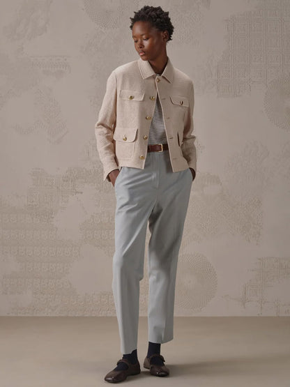 Indoors, a person stands with hands in pockets against a patterned wall wearing the Circolo 1901 Cropped 4-Pocket Jacket in Linen Bistretch, paired with a striped shirt, light blue trousers, dark socks, and loafers.