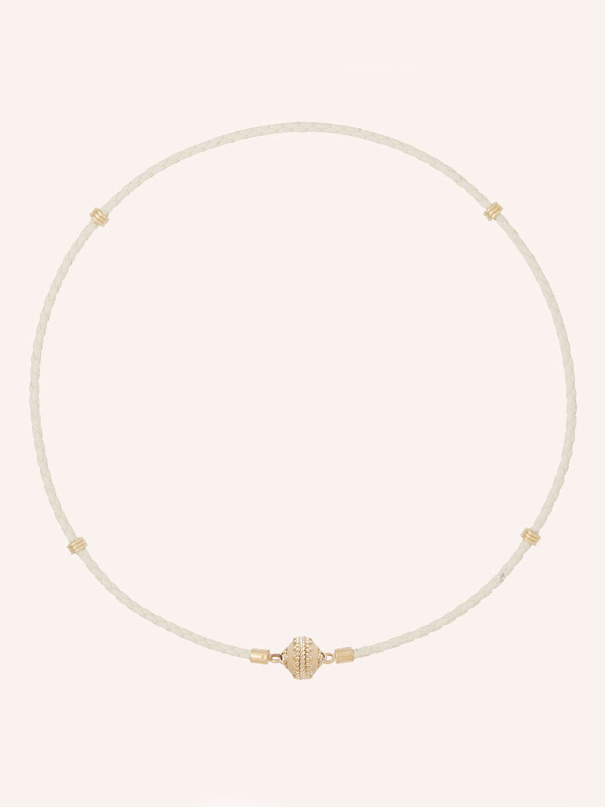The Clara Williams Aspen Braided Leather Cream Mini Necklace features hand-crafted braided cream genuine leather with subtle gold accents and a central gold bead, finished with a 14K yellow gold clasp, displayed on a plain light background.