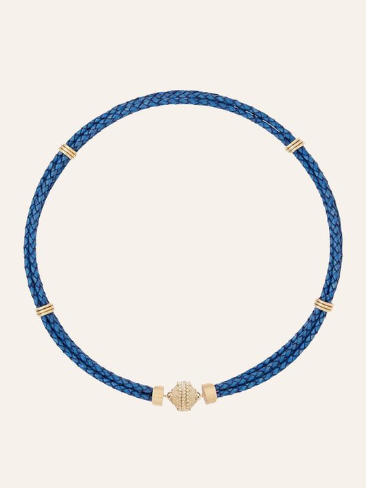 Clara Williams Aspen Braided Leather Denim Blue Necklace, crafted from braided denim blue genuine leather, features four 14K yellow gold bands and a central gold decorative bead, displayed against a plain light background.