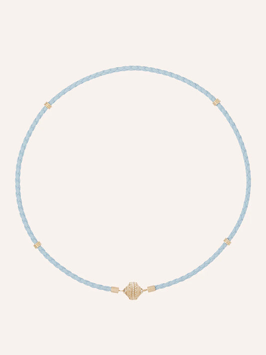 The Clara Williams Aspen Braided Light Blue Leather Mini Necklace features gold-tone bead accents and a central 14K yellow gold decorative bead, shown on a plain background.