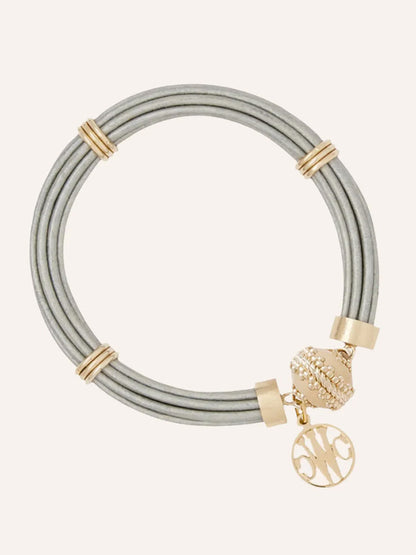 The Clara Williams Aspen Leather Platinum Bracelet features platinum leather strands, 14K yellow gold accents, a gold bead, and a round monogrammed charm hanging from its ClaraClasp.