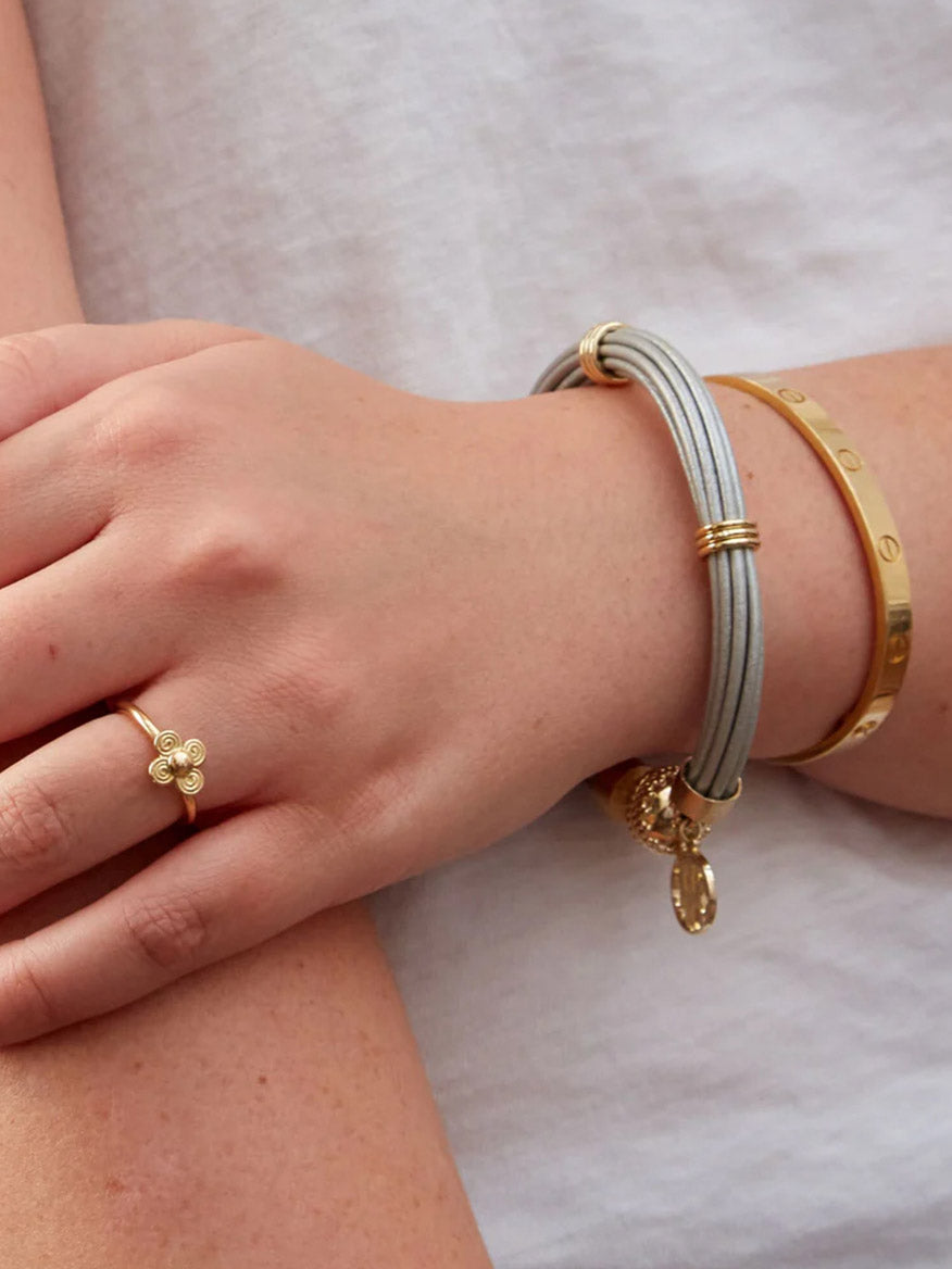 A hand wearing a 14K yellow gold clover ring, a Clara Williams Aspen Leather Platinum Bracelet, and a gold bangle set against a white shirt.