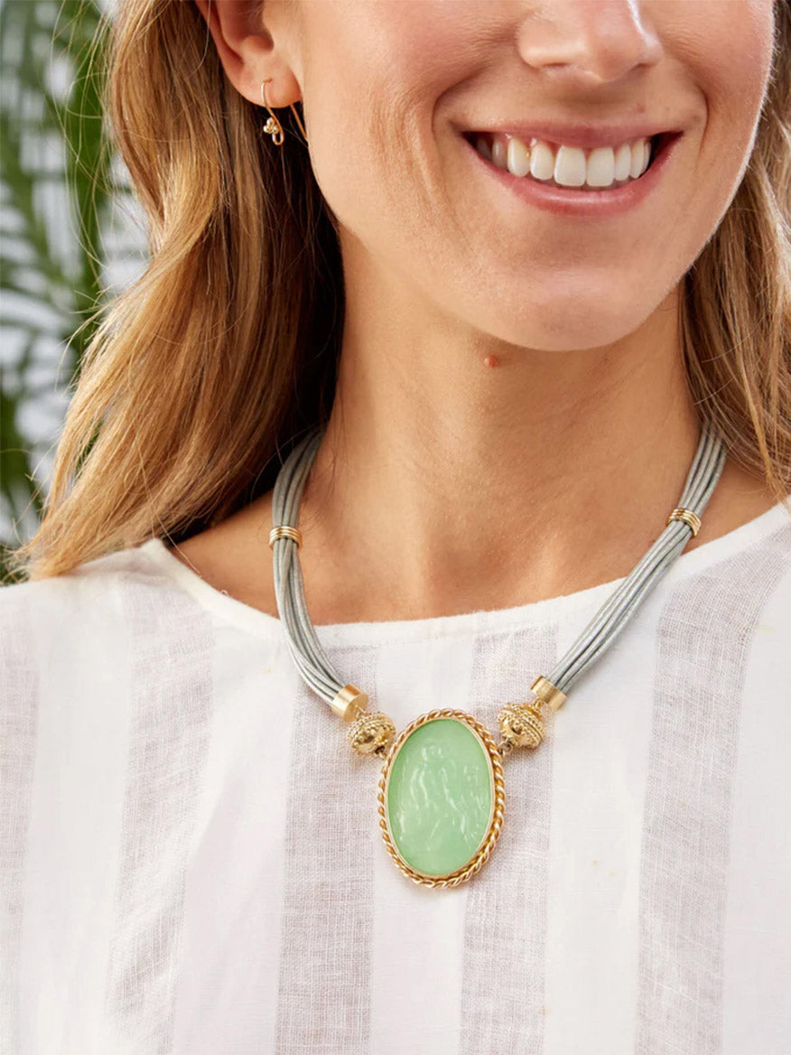 Woman wearing a white striped top and the Clara Williams Aspen Leather Platinum Silver Necklace with a large oval green pendant, 14K yellow gold accents, paired with small gold hoop earrings.