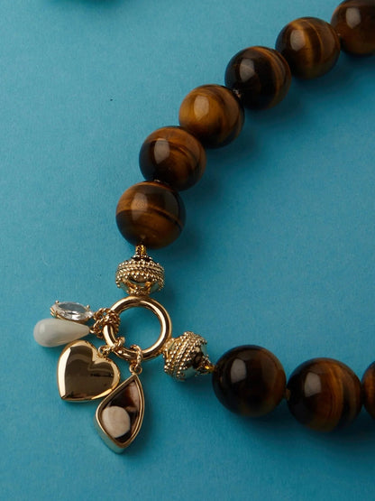 Clara Williams ClaraCharm™ Gold Ring Centerpiece: A brown beaded necklace with a 14K yellow gold signature clasp, adorned with three charms—a gold heart, a pearly teardrop, and a clear gemstone—shown against a blue background.
