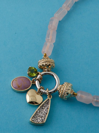 The Clara Williams ClaraCharm™ Rhodium Ring Mini Centerpiece is a beaded mini necklace with a central ring and five interchangeable charms—pink stone, green gem, textured silver, heart, and round bead—set on a light blue background.