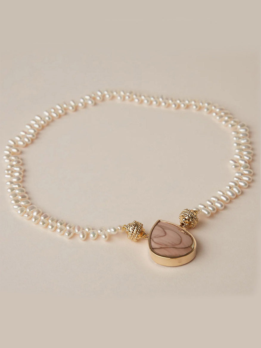 The Clara Williams Freshwater Drop Pearl Mini Necklace features freshwater pearls, gold beads, and a large teardrop-shaped brown stone pendant, elegantly displayed on a beige background.