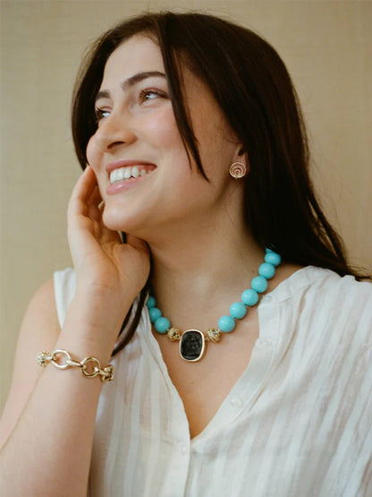 Woman with long dark hair smiles in a sleeveless white top, Clara Williams turquoise beaded necklace with black pendant, Gold Coast Bracelet in 14K yellow gold, and circular gold earrings against a neutral background.