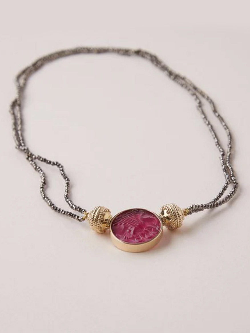 The Clara Williams Michel 2mm Coated Pyrite Double-Strand Mini Necklace features metallic strands, a round pink gemstone pendant, and two 14K yellow gold beads on either side, shown on a light background.