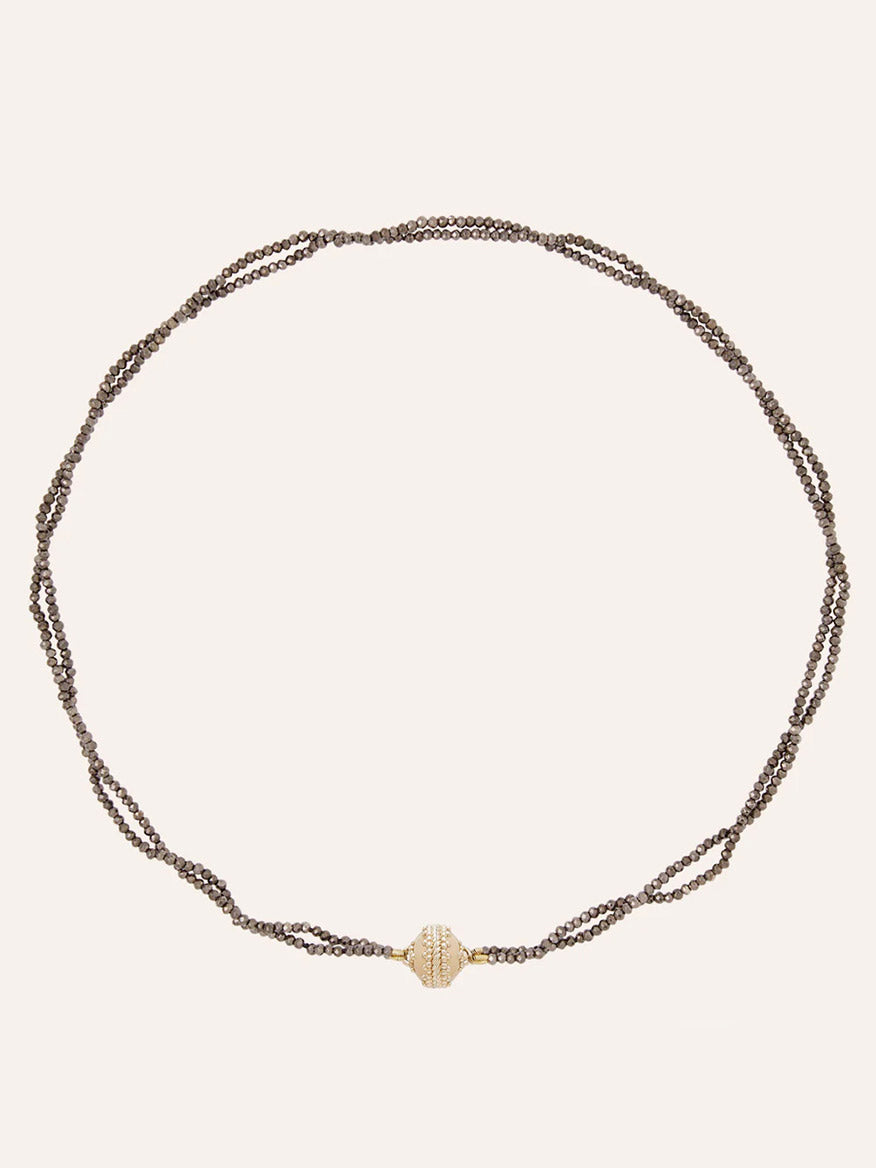 The Clara Williams Michel 2mm Coated Pyrite Double-Strand Mini Necklace features hand-strung brown chains and a central 14K yellow gold textured oval bead, elegantly displayed on a plain background.