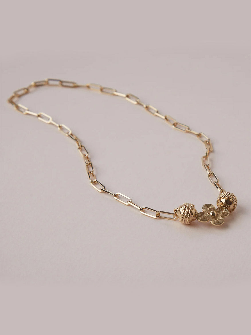 The Clara Williams Kingsbury Gold Mini Necklace features a 14K yellow gold paperclip chain with a ClaraClasp, two textured spherical beads, and a cluster of small round charms.