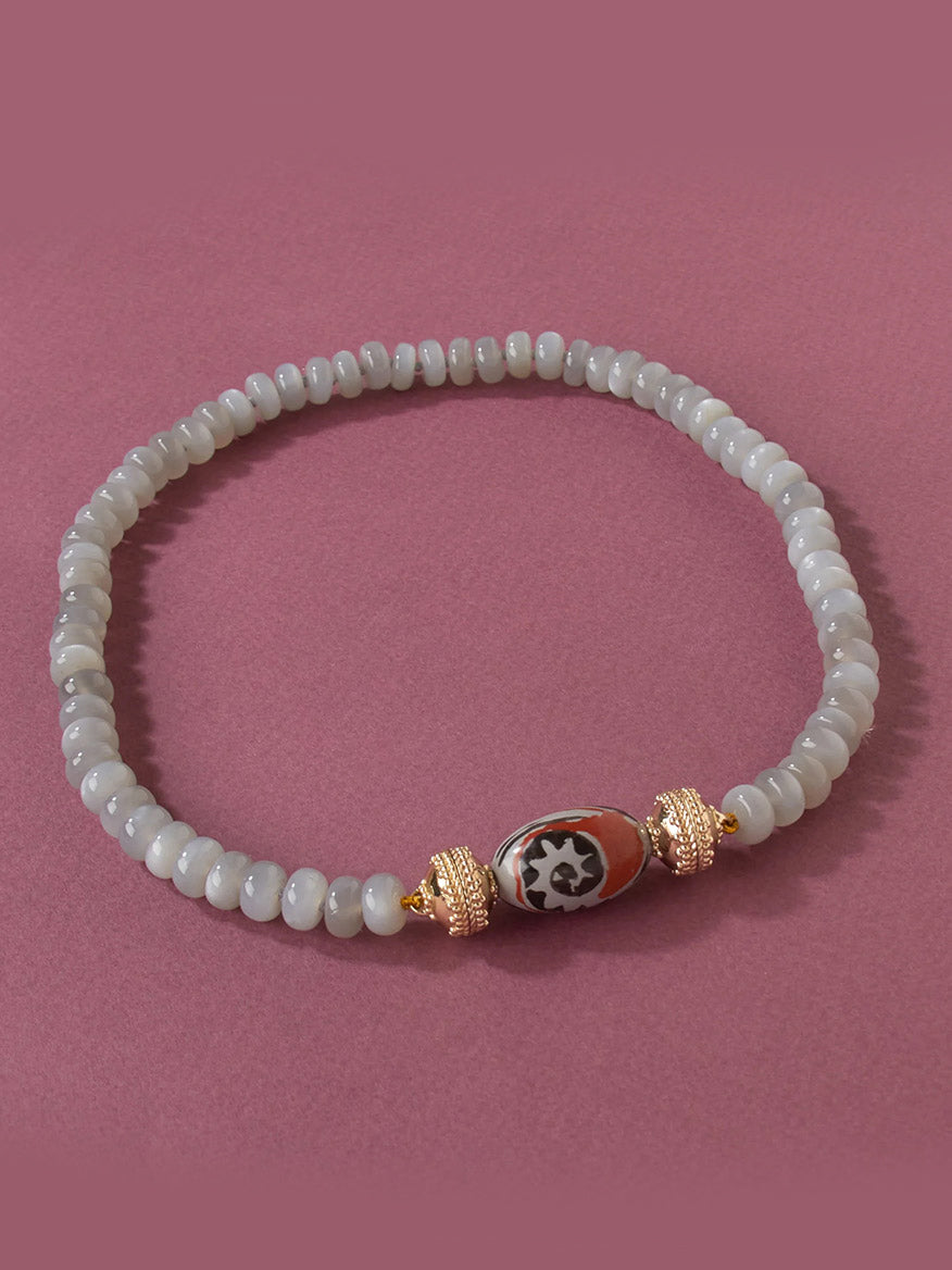 The Clara Williams Rust Horizon Ceramic Centerpiece features white round beads and a central patterned bead accented by two 14K yellow gold-toned details, finished with a sleek ceramic clasp and displayed on a pink background.