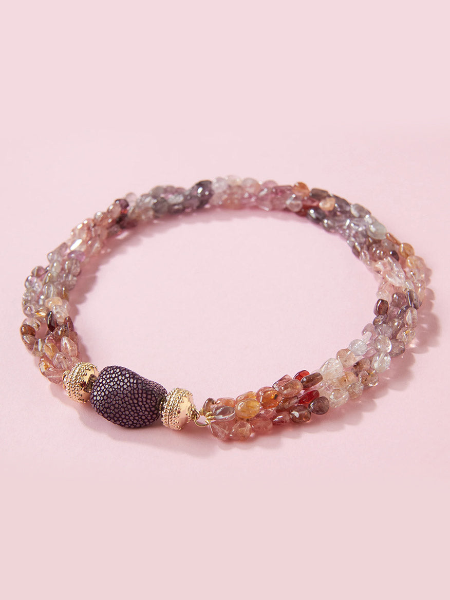 Showcasing the Clara Williams Small Plum Stingray Centerpiece, this multi-strand beaded bracelet features small, multicolored stones and a textured, dark purple centerpiece finished in a rich plum tone, set against a light pink background.
