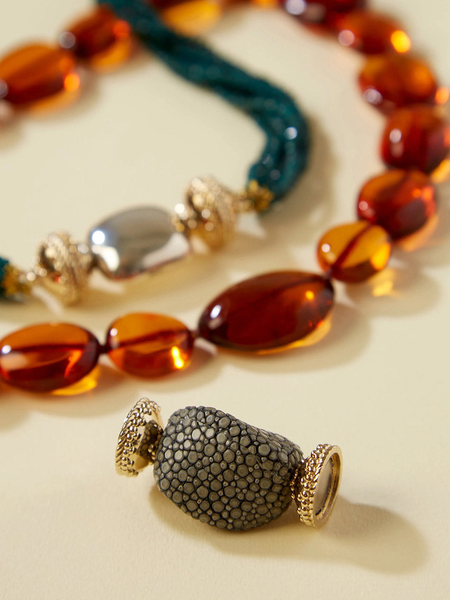Two beaded necklaces, one with amber-colored oval beads and another with green twisted beads, are laid on a pale surface. In the foreground is the Clara Williams Small Charcoal Stingray Centerpiece—a textured gray bead crafted from stingray hide and adorned with gold accents—adding an elegant touch that perfectly complements the 14K yellow gold clasp.