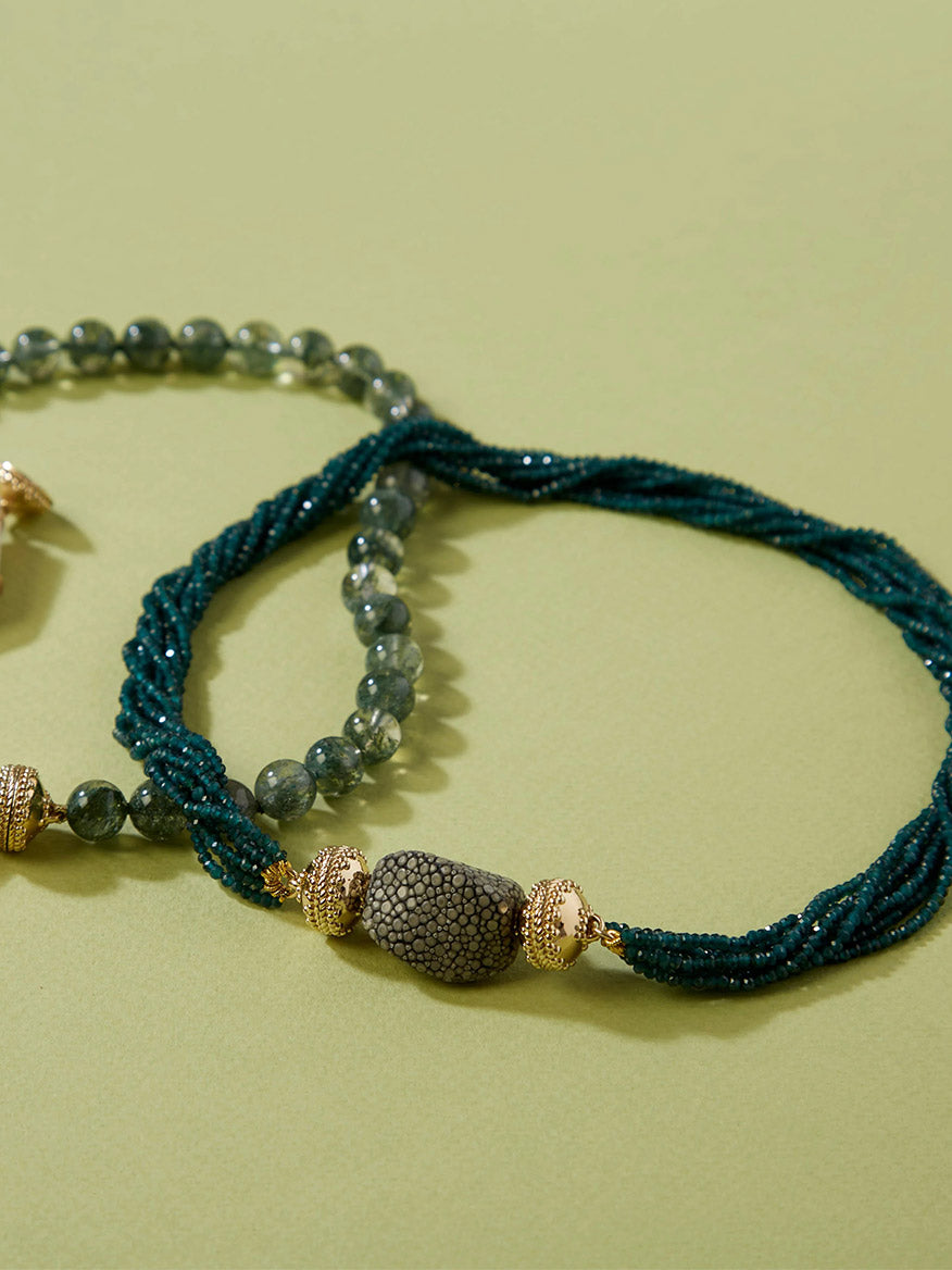 Two beaded necklaces on a light green surface: one with green round beads and gold accents finished with a 14K yellow gold clasp, the other featuring twisted dark green beads and the Clara Williams Small Charcoal Stingray Centerpiece, echoing the brand's signature style.