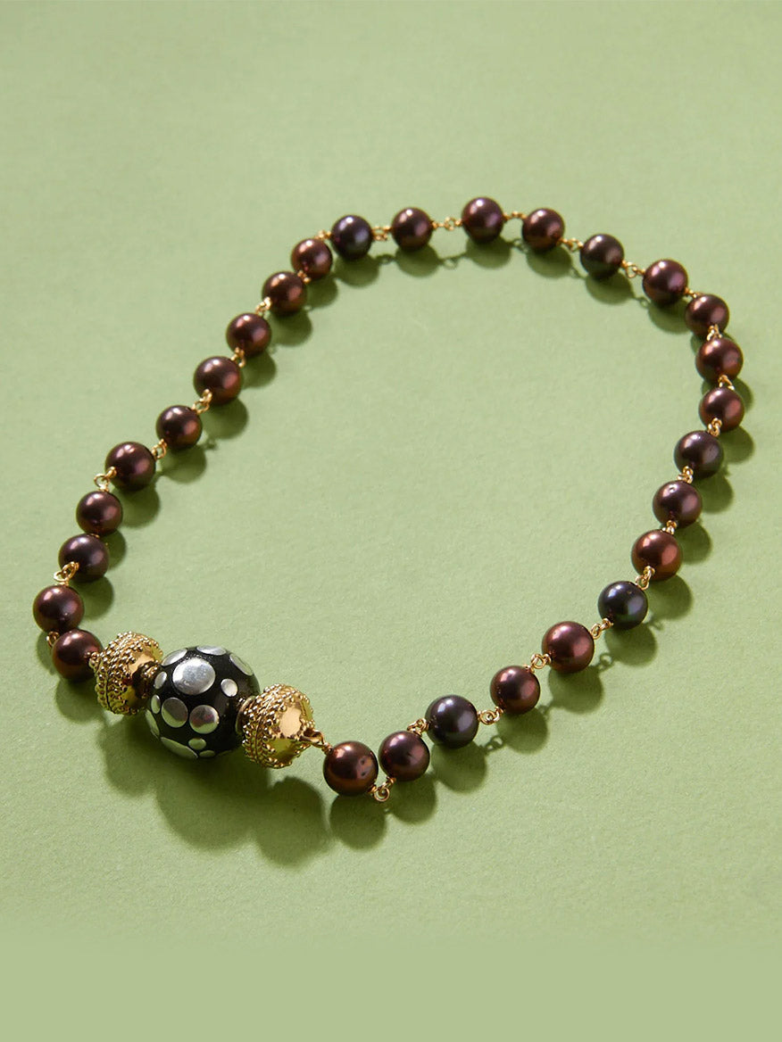 Clara Williams Sterling Silver Polka Dot Centerpiece necklace, with alternating dark purple and gold beads, showcases a large black enamel bead with white dots at the center, displayed on a light green background.
