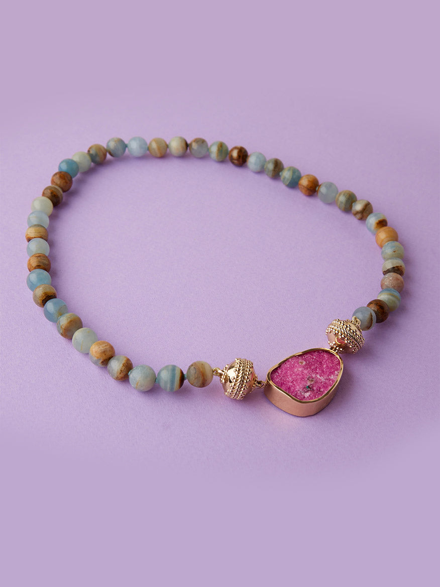 Clara Williams Victoire 10mm Blue Calcite Necklace featuring round blue calcite beads and a pink gemstone pendant accented with gold details, finished with a 14K yellow gold clasp, displayed on a light purple background.