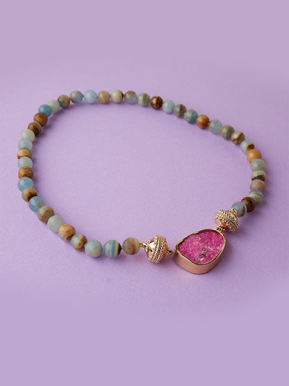 Clara Williams Victoire 10mm Blue Calcite Necklace featuring round blue calcite beads and a pink gemstone pendant accented with gold details, finished with a 14K yellow gold clasp, displayed on a light purple background.