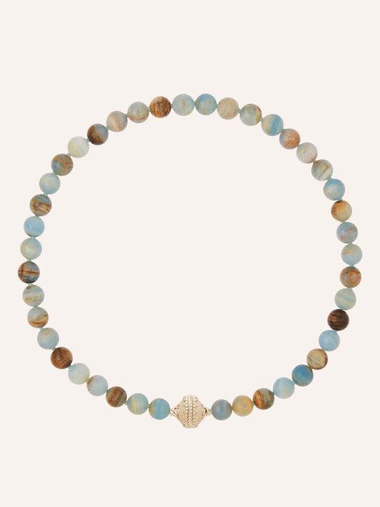 The Clara Williams Victoire 10mm Blue Calcite Necklace features round, blue and brown marbled natural calcite stones and a 14K yellow gold clasp, displayed in a circular arrangement on a plain background.