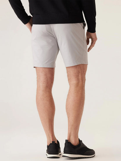 Photographed from behind, a person is standing, wearing a black long-sleeve shirt, DEKE Galaxy Bermuda Ultra 4X Stretch Twill shorts in Stone, and black sneakers.