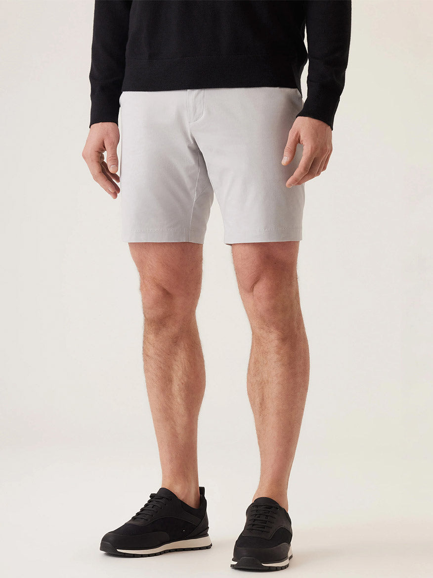 A person wears a black long-sleeve top, DEKE Galaxy Bermuda Ultra 4X Stretch Twill shorts in Stone, and black sneakers while standing against a light background.
