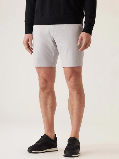 A person wears a black long-sleeve top, DEKE Galaxy Bermuda Ultra 4X Stretch Twill shorts in Stone, and black sneakers while standing against a light background.