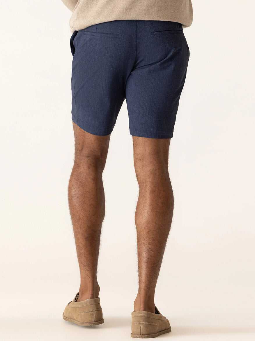A person stands facing away, wearing DEKE Galaxy Bermuda Nano Nylon Warp Knit in Indigo shorts for wrinkle resistance and UV protection, paired with a beige top and tan loafers against a plain background.