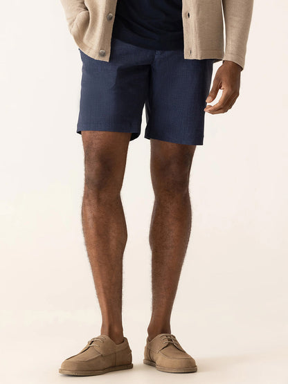 A man stands against a plain background wearing an indigo DEKE Galaxy Bermuda Nano Nylon Warp Knit, navy shorts, a dark shirt, and tan shoes.