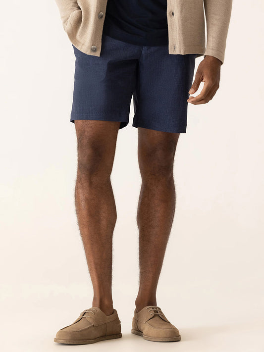 A man stands against a plain background wearing an indigo DEKE Galaxy Bermuda Nano Nylon Warp Knit, navy shorts, a dark shirt, and tan shoes.