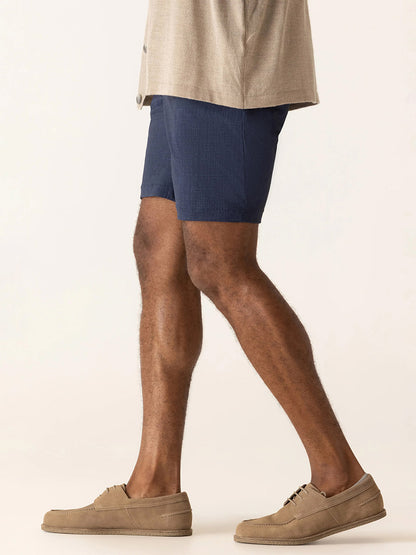 A person wearing a beige shirt, DEKE Galaxy Bermuda Nano Nylon Warp Knit shorts in Indigo, and beige loafers is shown walking, photographed from the shoulders down against a plain background.