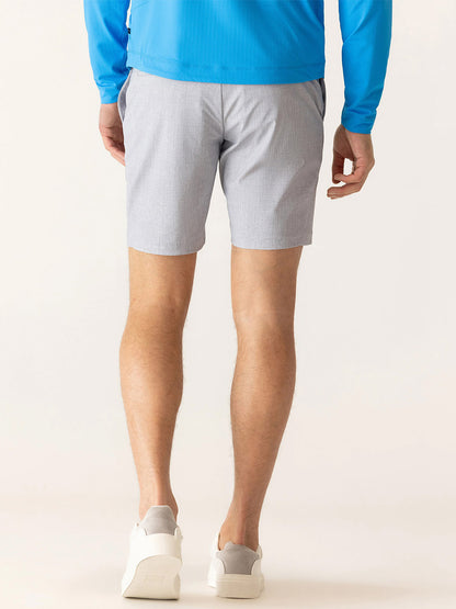A person stands facing away, wearing a blue long-sleeve shirt, DEKE Galaxy Bermuda Nano Nylon Warp Knit shorts in Silver, and white sneakers against a neutral background.