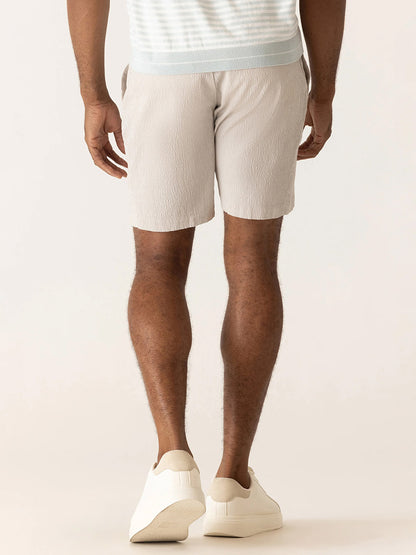 A man stands facing away, wearing the DEKE Galaxy Bermuda Supernatural Seersucker in Sand, paired with a striped shirt and white sneakers—effortlessly capturing refined summer style against a neutral background.