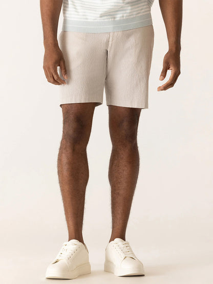 A man showcases effortless summer style in DEKE Galaxy Bermuda Supernatural Seersucker shorts in Sand, paired with a striped shirt and white sneakers, against a plain background.