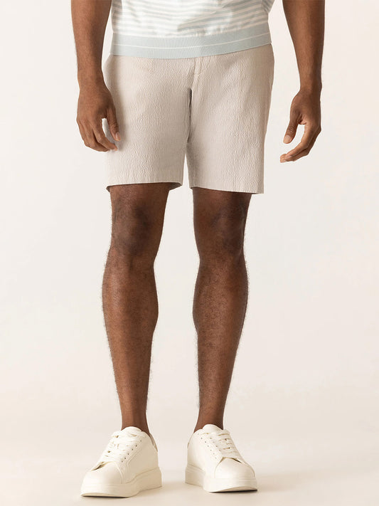 A man showcases effortless summer style in DEKE Galaxy Bermuda Supernatural Seersucker shorts in Sand, paired with a striped shirt and white sneakers, against a plain background.