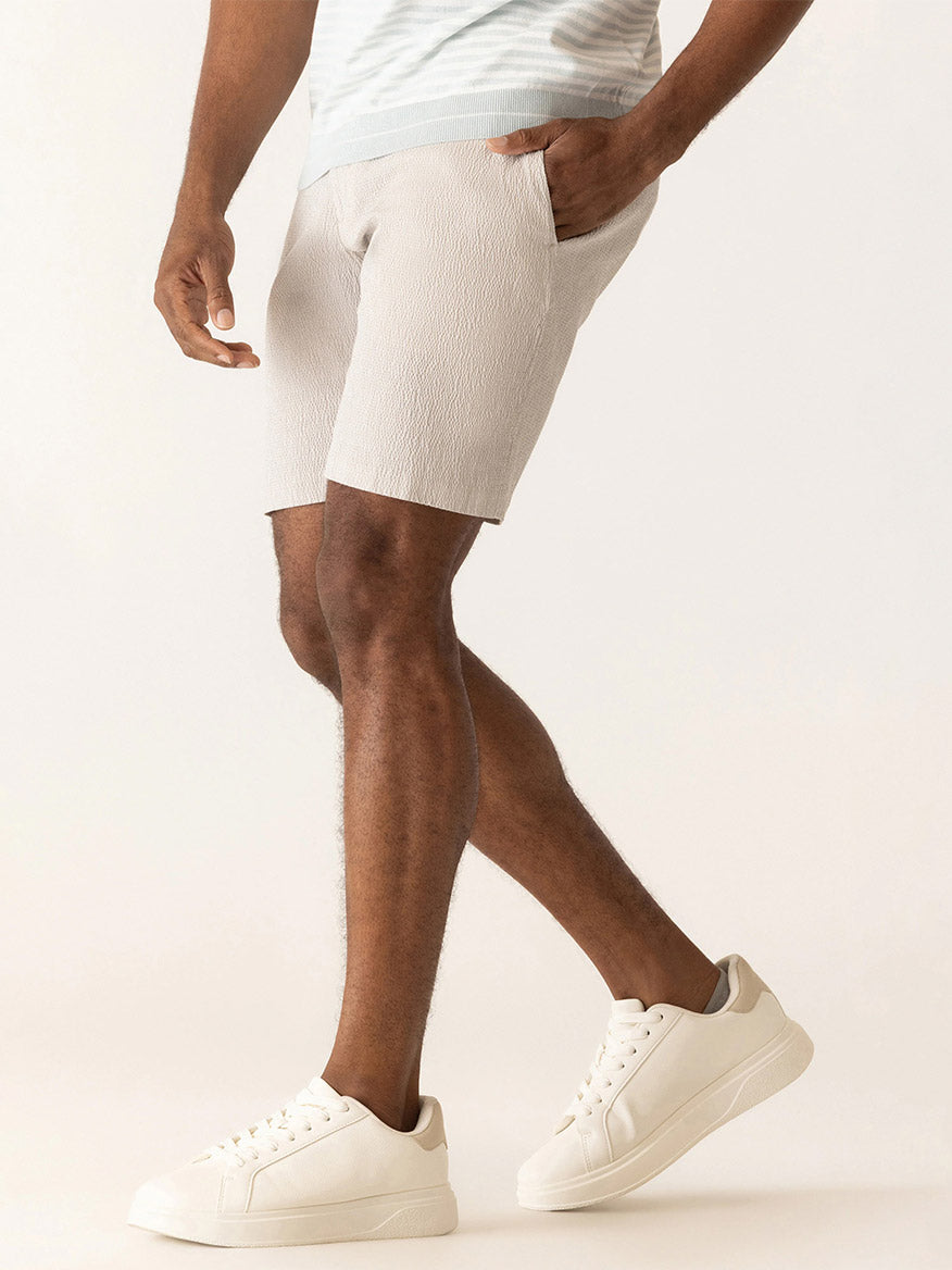 A man in a light blue shirt, DEKE Galaxy Bermuda Supernatural Seersucker shorts in Sand, and white sneakers stands with one hand in his pocket against a plain background—effortlessly capturing summer style.