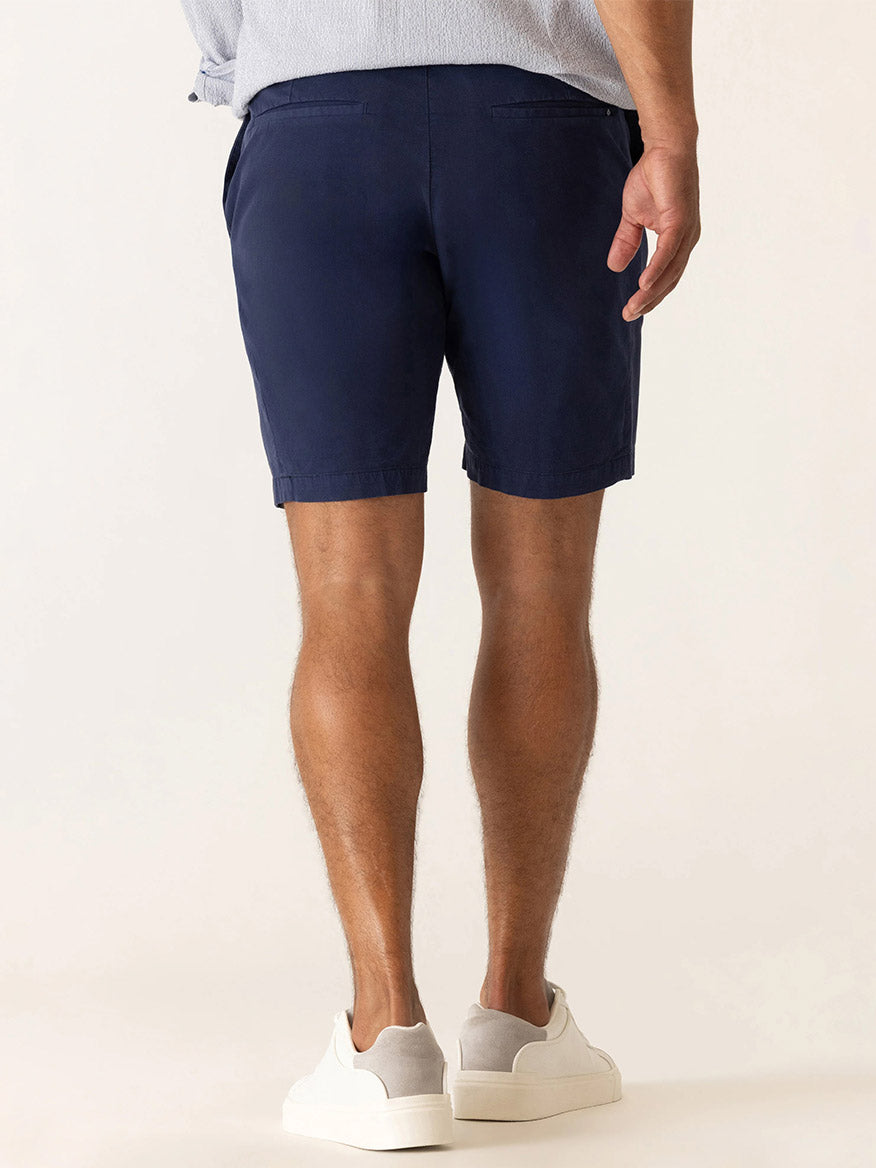 A man faces away, wearing a light blue shirt, white sneakers, and DEKE Galaxy Bermuda Ultra 4X Stretch Twill shorts in Indigo with a tailored fit against a plain background.