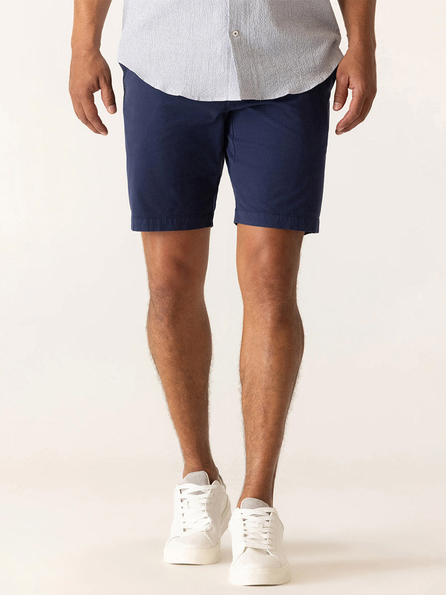 A man in a light button-up shirt, DEKE Galaxy Bermuda Ultra 4X Stretch Twill shorts in Indigo with a tailored fit, and white sneakers stands against a plain background.