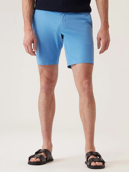 Wearing DEKE Galaxy Bermuda Ultra 4X Stretch Twill in Lake Blue, a dark top, and black sandals, this person stands against a plain light background, effortlessly showcasing performance-driven sophistication.