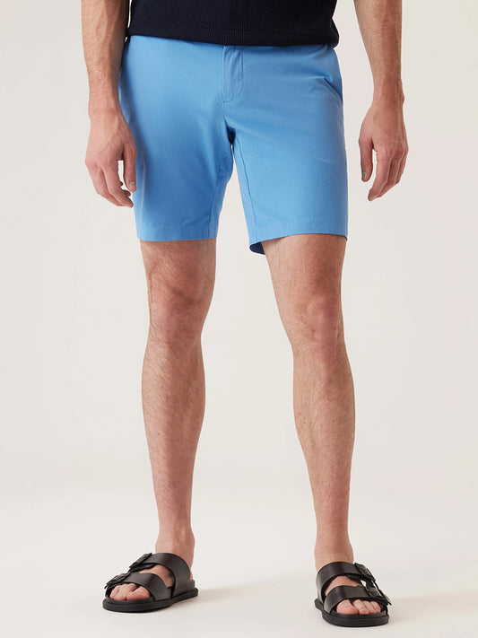 Wearing DEKE Galaxy Bermuda Ultra 4X Stretch Twill in Lake Blue, a dark top, and black sandals, this person stands against a plain light background, effortlessly showcasing performance-driven sophistication.