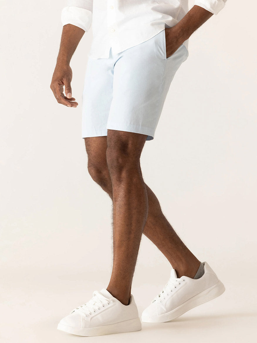 A person wearing a white shirt, DEKE Galaxy Bermuda Ultra 4X Stretch Twill in Ice Blue shorts, and white sneakers stands against a plain light background.