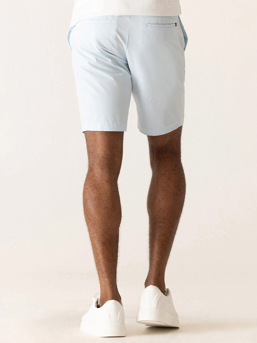 A person is shown from the back, wearing DEKE Galaxy Bermuda Ultra 4X Stretch Twill shorts in Ice Blue and white sneakers, standing against a neutral background.