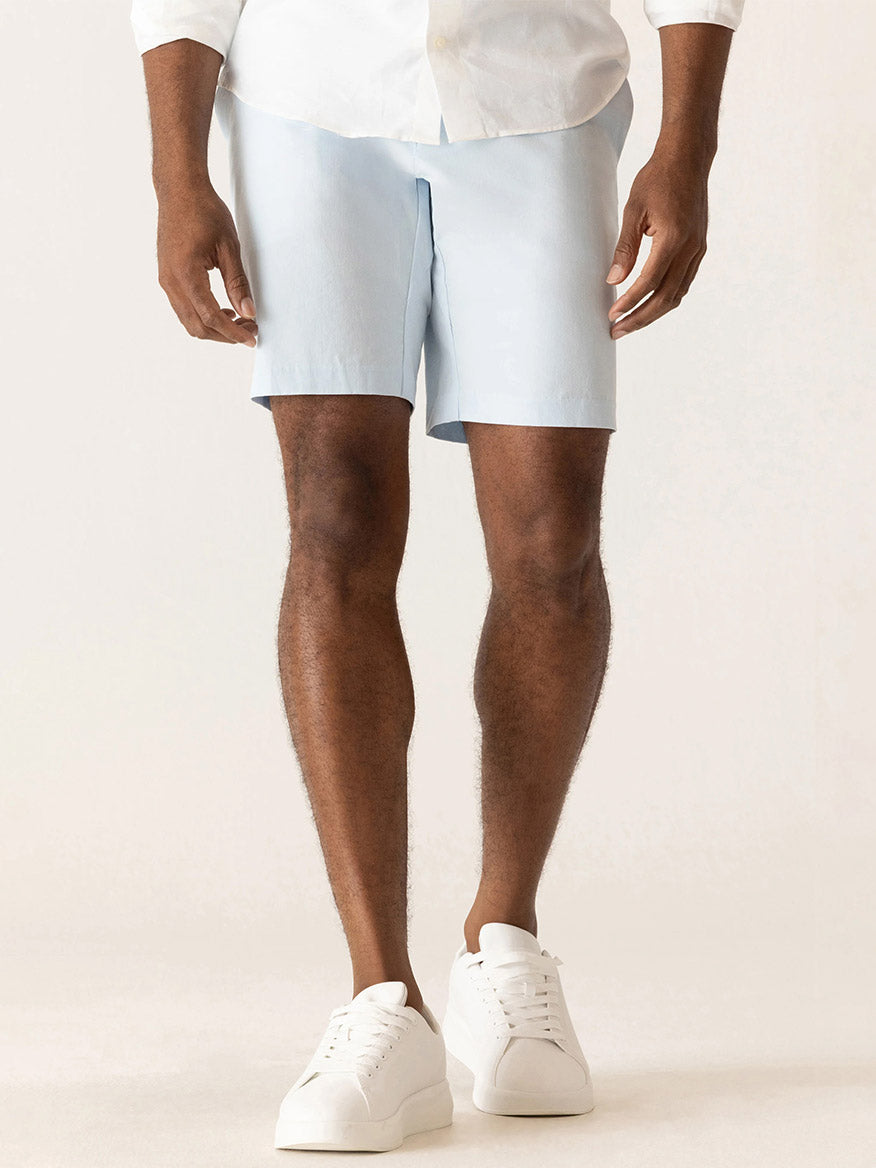 A person wearing a white shirt, DEKE Galaxy Bermuda Ultra 4X Stretch Twill in Ice Blue shorts, and white sneakers stands against a plain light background.