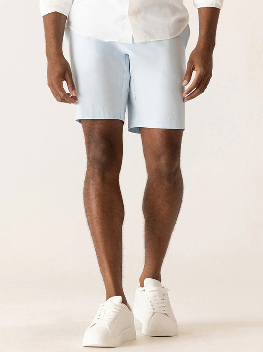 A person wearing a white shirt, DEKE Galaxy Bermuda Ultra 4X Stretch Twill in Ice Blue shorts, and white sneakers stands against a plain light background.