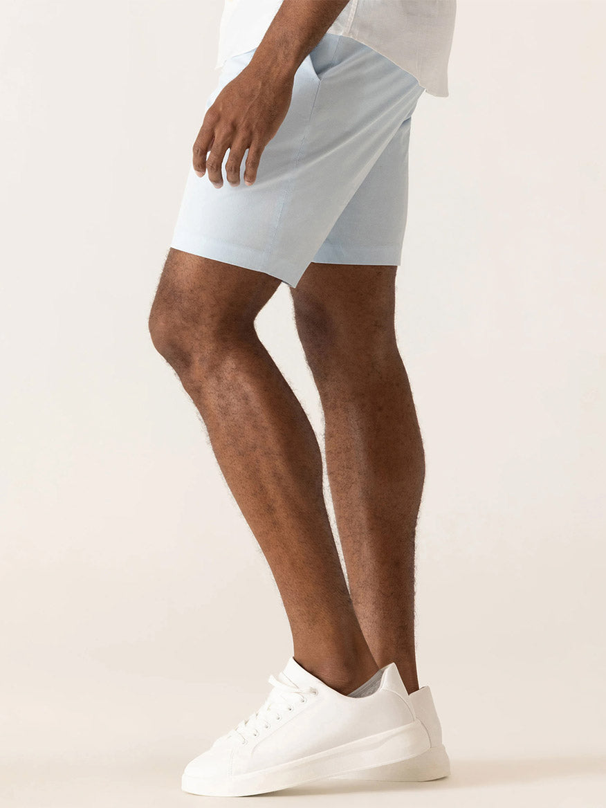 A man stands sideways against a plain light background wearing DEKE Galaxy Bermuda Ultra 4X Stretch Twill shorts in Ice Blue, paired with a white shirt and white sneakers.