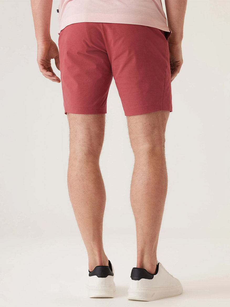 A person faces away, wearing the DEKE Galaxy Bermuda Ultra 4X Stretch Twill in Nantucket Red and a light pink shirt, paired with white sneakers with black details against a plain light background.