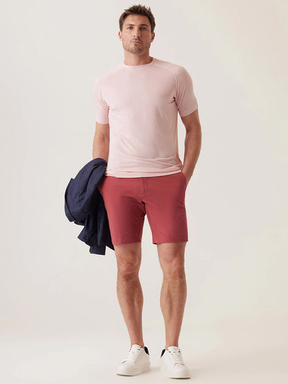 A man stands in a studio wearing a light pink t-shirt, white sneakers, and DEKE Galaxy Bermuda Ultra 4X Stretch Twill shorts in Nantucket Red, holding a navy jacket in his left hand.