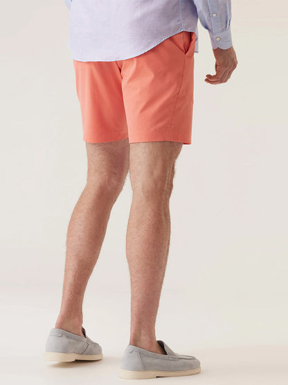 A man stands facing away, wearing a light blue shirt and DEKE Galaxy Bermuda Ultra 4X Stretch Twill in Sunset shorts with a tailored fit, paired with light gray loafers against a plain background.