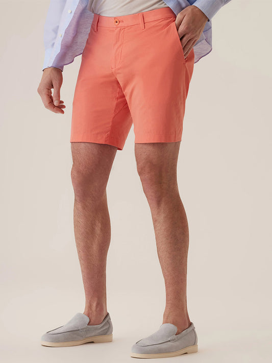 A person wearing DEKE Galaxy Bermuda Ultra 4X Stretch Twill in Sunset, a light blue shirt, and light gray loafers stands with one hand in their pocket, achieving a stylish, tailored look that's also performance-driven.